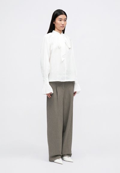 Biała bluzka z krawatem przy szyi i balonowymi rękawami, noszona z szerokimi szarymi spodniami. Modelka ma na sobie białe, spiczaste szpilki. Minimalistyczny design.