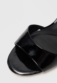 Nærbilde av den fremre delen av en glanset svart åpen tå-sandal med en bred stropp over foten mot en ensfarget bakgrunn.