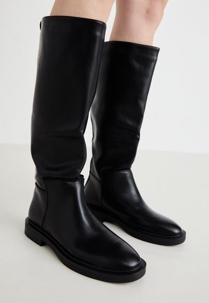 Bottes en cuir noires montant jusqu'aux genoux portées sur des jambes nues, avec des bouts arrondis et des semelles plates en caoutchouc sur un fond clair.