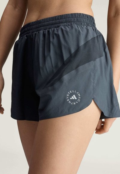 Nahaufnahme einer Person, die dunkelgraue Sportshorts mit elastischem Bund und einem kreisförmigen Adidas by Stella McCartney Logo trägt.