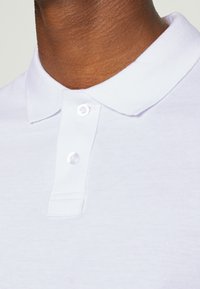 Hvit polo-skjorte med klassisk krage, to klare plastknapper og teksturert strikket stoff. Nærbilde av halsområdet.