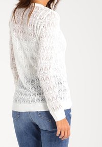 Mujer con un suéter de punto de encaje de manga larga blanca y jeans azules, vista desde atrás y de lado, contra un fondo liso.