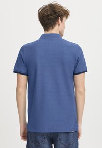 Blå polo skjorte med ribbet krage og mansjetter, teksturert stoff, sidespalter i sømmen, og en liten merkelapp nederst.