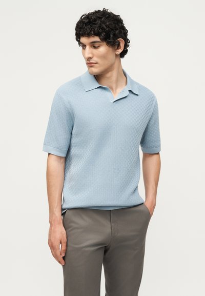 Άνδρας που φοράει ανοιχτό μπλε polo με κοντά μανίκια και υφή, και γκρι παντελόνι, κοιτάζει αριστερά με το ένα χέρι στην τσέπη, σε λευκό φόντο.