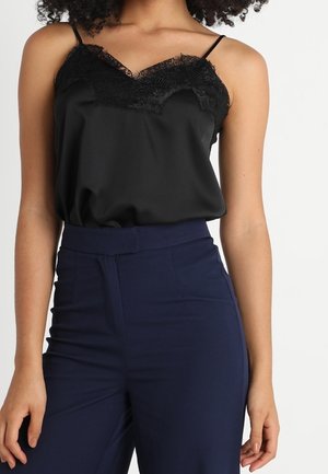 Kobieta ubrana w czarną koszulkę na ramiączkach z koronkową lamówką, schowaną w granatowe spodnie z wysokim stanem, na jednolitym tle.