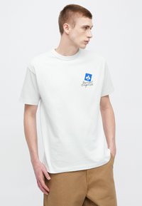 Biele bavlnené tričko s krátkymi rukávmi, okrúhlym výstrihom a modrým emblémom na ľavej strane hrudníka s nápisom "the new originals."