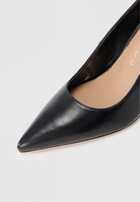 Escarpin en cuir noir à talon haut avec bout pointu et texture lisse et élégante. L'intérieur comprend une semelle beige.