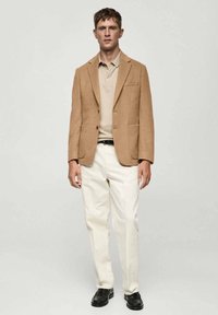 Smeđi blejzer s izrezanim reverima i dva prednja džepa, uparen s bež polo majicom, krem hlačama i crnim cipelama. Uočljive glatke teksture.