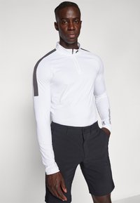 Hvit langermet topp med kvart glidelås og grå detaljer på skuldrene, sammen med mørkegrå shorts med knappeåpning.