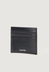 Melnas ādas kartes turētājs ar vairākiem kartēm paredzētiem nodalījumiem un baltu "Calvin Klein" logo apakšā. Gluda tekstūra ar tīrām līnijām.