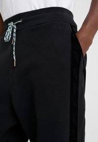 Pantalones de chándal negros hechos de suave algodón con una cinturilla acanalada, que presentan un cordón trenzado azul y blanco y acentos de terciopelo en los laterales.