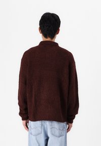 Maglione marrone in tessuto strutturato con un fit leggermente corto, maniche lunghe e colletto. Indossato sopra jeans in denim blu chiaro.