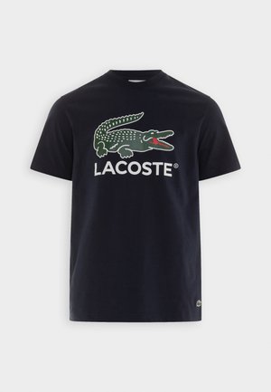 T-shirt en coton bleu marine avec un grand logo crocodile vert et le texte "LACOSTE" en blanc, coupe classique à col rond. Texture douce.