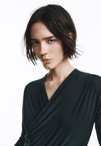 Čierny zavinovací top z jemnej látky s hlbokým výstrihom do V, dlhými rukávmi a zdobeným dizajnom, ktorý zvýrazňuje hladkú textúru a priliehavý strih.