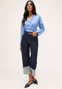 Pantaloni din denim cu un ton închis, prezentând un tiv unic pliat cu un detaliu din denim deschis. Asortați cu o cămașă cu nasturi de culoare albastră deschisă cu dungi.