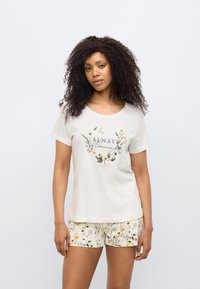 Μπεζ βαμβακερό t-shirt με στρογγυλή λαιμόκοψη, διακοσμημένο με λουλουδάτο σχέδιο και το κείμενο "Πάντα ανθισμένο." Συνδυάζεται με λευκά σορτς διακοσμημένα με κίτρινο λουλουδάτο μοτίβο.