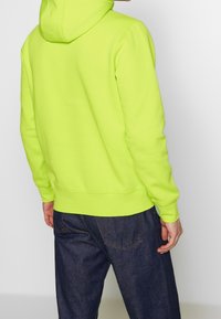 Felpa con cappuccio verde neon, maniche lunghe, polsini e orlo ribattuti, abbinata a jeans in denim scuro, vista dalla schiena.