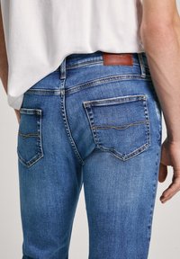 Jeans de mezclilla azul con un corte ajustado, que cuentan con un detalle cosido en los bolsillos traseros y un parche de cuero marrón con el logo en la cintura.