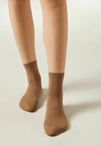 Calcetines de algodón marrón con mezcla, con un puño acanalado y textura suave, que se ajustan justo por encima del tobillo, usados sobre piernas desnudas.