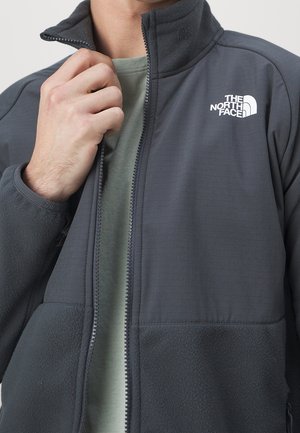 Mężczyzna w ciemnoszarej kurtce North Face na zamek błyskawiczny na jasnooliwkowej koszuli, trzyma za kołnierz kurtki lewą ręką.