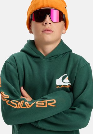 Zelená mikina s kapucí s velkým bílým a oranžovým logem Quiksilver, doplněná oranžovou pletenou čepicí a reflexními slunečními brýlemi s růžovými skly.