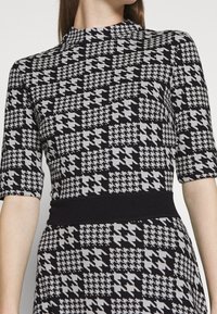 Robe en tricot avec un motif pied-de-poule noir et gris, manches courtes, col haut et une ceinture noire pour souligner la taille.
