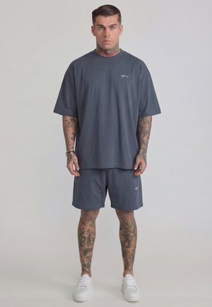 Mann mit Tattoos, der ein dunkelgraues oversized T-Shirt und passende Shorts trägt, steht vor einem schlichten hellen Hintergrund mit weißen Sneakers.