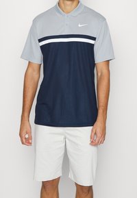 Pánská polo košile Nike s dvoubarevným designem, světle šedými rukávy a námořnickým modrým tělem, doplněná bílým horizontálním pruhem. Oblečeno s bílými šortkami.