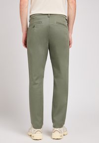 Smal passform i olivgröna byxor med en slät textur, som har bakfickor och en knappdetalj, tillsammans med beigea träningsskor.