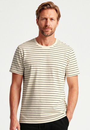 Mann mit hellbraunen Haaren und Bart, der ein cremefarbenes T-Shirt mit dünnen schwarzen horizontalen Streifen trägt und vor einem schlichten Hintergrund steht.