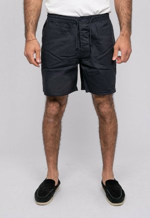 Homme portant un short noir à cordon, un t-shirt blanc et des chaussures noires sans lacets, debout sur un fond clair uni.