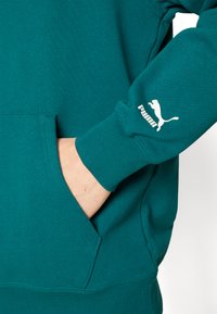 Tyrkysová mikina s kapucňou vyrobená z jemného materiálu, s klokanie vreckom, rebrovanými manžetami a bielym logom Puma na rukáve.