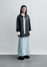 Camisa oversized em couro preto, camisa branca de botão por baixo, saia maxi de ganga azul-claro e chinelos pretos felpudos. Texturas suaves em todo o conjunto.