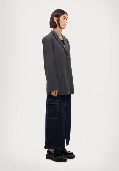 Γκρίζα oversized σακάκι, τζιν παντελόνι σε φαρδιά γραμμή με τσέπες και μαύρα χοντρά παπούτσια. Το μοντέλο στέκεται σε ένα λευκό φόντο.