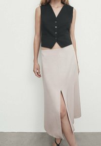 Black, sleeveless vest with a V-neck and five buttons paired with a beige, knee-length skirt featuring a front slit. Smooth fabric texture.