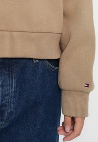 Beige genser med ribbede mansjetter, kombinert med mørkeblå denimjeans. Merkbar logo-detalj på mansjetten. Glatt tekstur og avslappet passform.