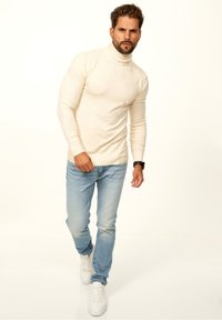 Beiger Rollkragenpullover aus einem weichen Material, kombiniert mit hellblauen Jeans und weißen Sneakers, im schmalen Schnitt.