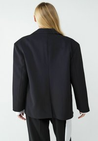 Czarny dopasowany żakiet o zdefiniowanej sylwetce, z klapami i pojedynczym rozcięciem z tyłu. Gładka tkanina z minimalnym zdobieniem.