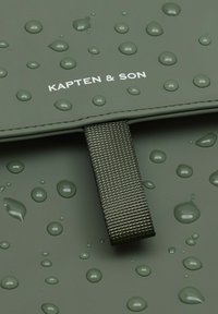 Grijs-groene waterdichte tas met waterdruppels en een geweven riem; bevat het "KAPTEN & SON" logo in het wit op het oppervlak.