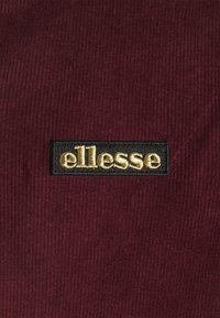 Bordeaux corduroy stof met een zwart rechthoekig label met het geborduurde logo "ellesse" in goud. Textuur lijnen zichtbaar in de stof.