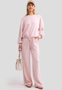 Chándal rosa que presenta una parte superior de corte relajado con cintura ajustable y pantalones de pierna ancha. Textura suave combinada con un bolso de color claro.