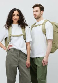 Två personer står bredvid varandra, klädda i vita t-shirts. Den ena har olivgröna byxor, medan den andra har ljusgrön byxor. Båda bär khaki ryggsäckar.