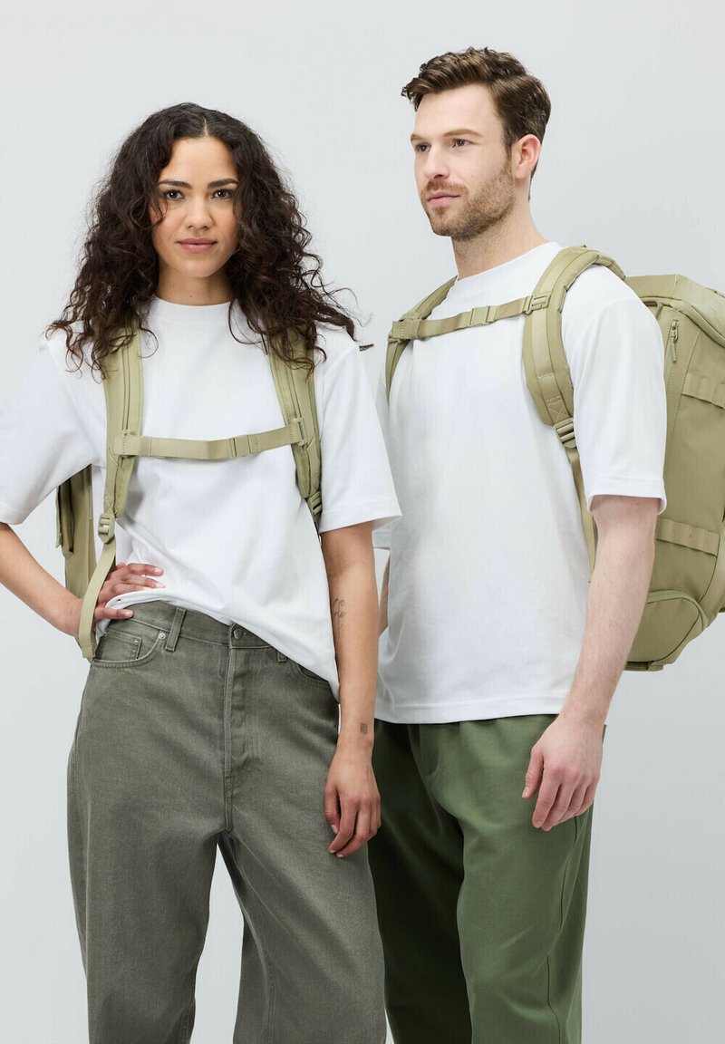 Två personer står bredvid varandra, klädda i vita t-shirts. Den ena har olivgröna byxor, medan den andra har ljusgrön byxor. Båda bär khaki ryggsäckar.