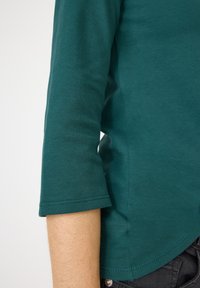 Türkisfarbenes Langarmshirt mit abgerundetem Saum, engen Ärmeln und weichem Material, seitlich gezeigt mit einem sichtbaren Arm.