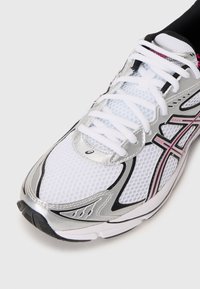 Scarpa sportiva bianca con tomaia in tessuto a rete, dettagli argento, decorazioni nere e rosa, e suola testurizzata per una presa migliore.