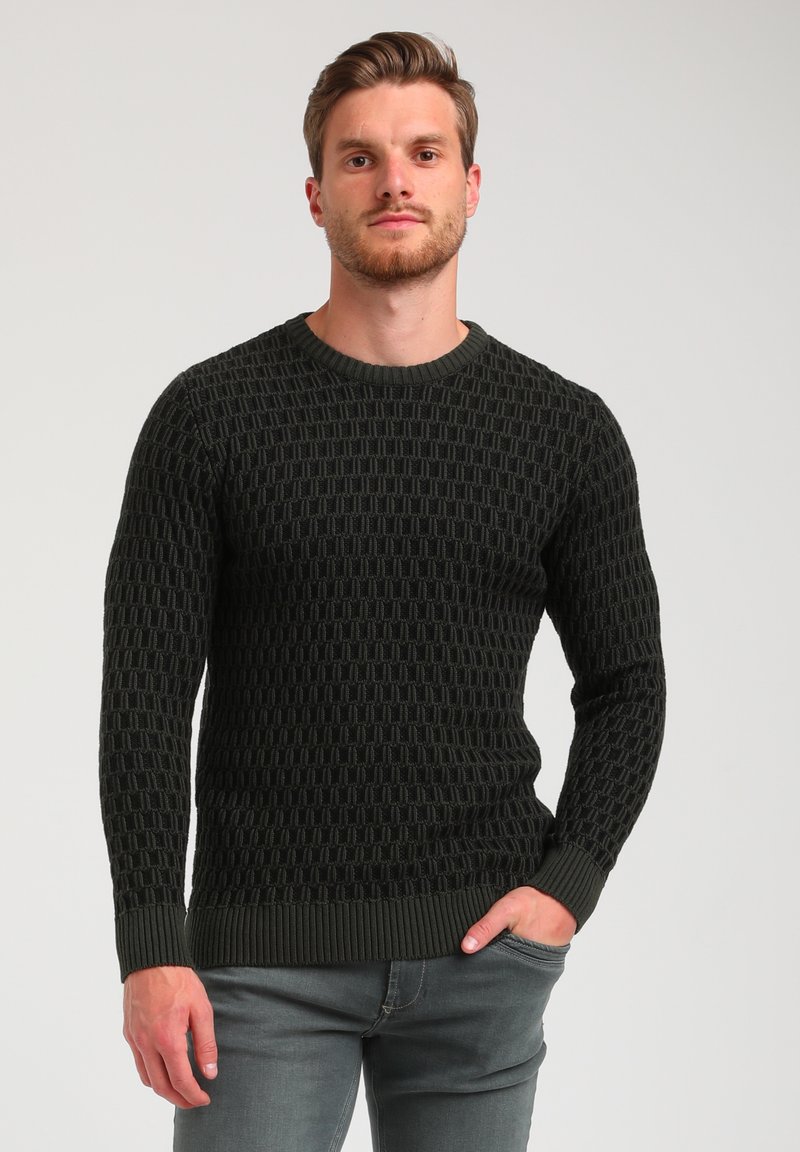 Suéter de lana de color verde oscuro con un patrón texturizado, escote redondo y puños acanalados. Combinado con unos jeans de color gris claro.
