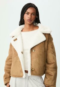 Donna con capelli intrecciati che indossa orecchini a cerchio dorati, un giubbotto di pelliccia scamosciata color beige sopra una maglietta bianca e pantaloni grigio chiaro, su uno sfondo semplice e chiaro.