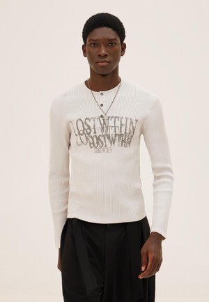 Jonge man die een witte geribbelde lange-ärmelig shirt draagt met de gelaagde tekst "LOST WITHIN" en zwarte broek, staand tegen een effen achtergrond.