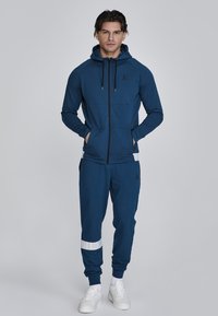 Navyblaues Zip-Hoodie mit Kordelzügen, Fronttaschen und weißen Akzenten. Kombiniert mit passenden Jogginghosen mit einem weißen Streifen.