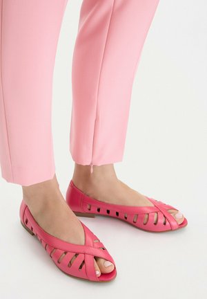 Pies con zapatos planos abiertos de color rosa con detalles recortados, combinados con pantalones de longitud hasta el tobillo en rosa claro, que cuentan con una pequeña cremallera lateral.